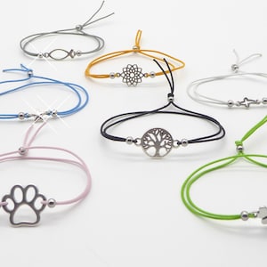 Könnte beinhalten: Ein Set aus sieben bunten Armbändern mit verschiedenen silbernen Anhängern. Die Anhänger umfassen einen Fisch, eine Blume, einen Baum, ein Kreuz, einen Pfotenabdruck und ein Kleeblatt.
