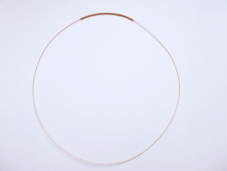 May include: A delicate necklace featuring a thin, silver-colored chain with a small, copper-colored accent. The necklace is circular in shape, resting against a white background. The minimalist design suggests a focus on simplicity and elegance.