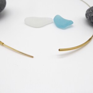 May include: A gold-colored necklace with a minimalist design. The necklace is open, showing the two ends of the curved bar. The background includes a gray stone, and two pieces of sea glass in white and blue.