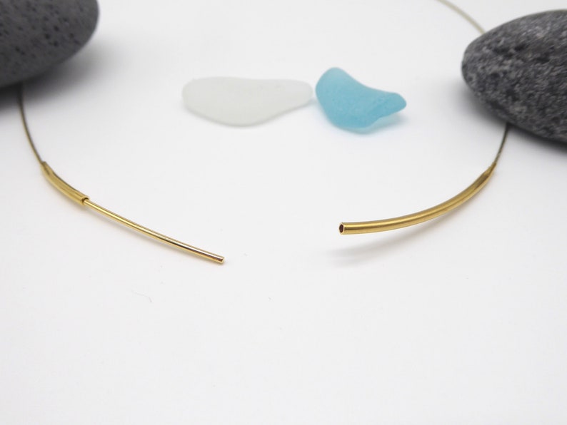 May include: A gold-colored necklace with a minimalist design. The necklace is open, showing the two ends of the curved bar. The background includes a gray stone, and two pieces of sea glass in white and blue.