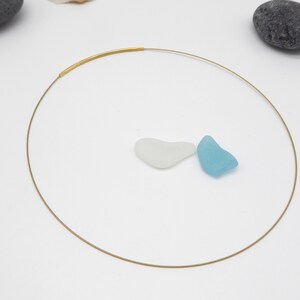 May include: A delicate gold-tone choker necklace with a minimalist design. The necklace features a thin, circular wire with a thicker, textured gold bar at the front. Two pieces of sea glass, one white and one light blue, are placed near the necklace.