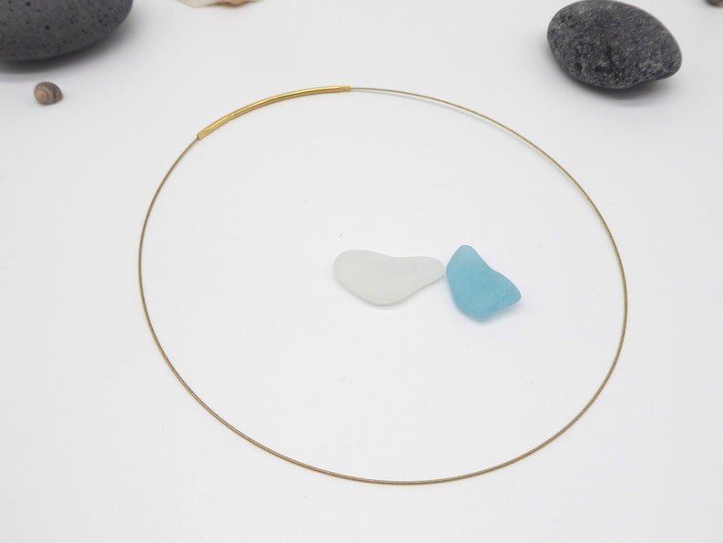May include: A delicate gold-tone choker necklace with a minimalist design. The necklace features a thin, circular wire with a thicker, textured gold bar at the front. Two pieces of sea glass, one white and one light blue, are placed near the necklace.
