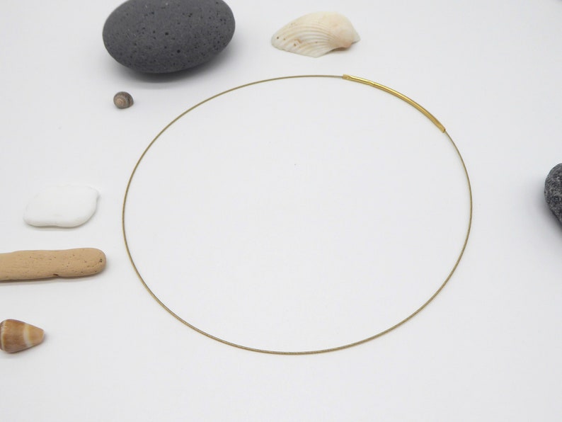 May include: A delicate gold-tone necklace with a minimalist design. The necklace features a thin, circular wire with a thicker, curved gold bar at the front. The background includes natural elements like stones and shells.