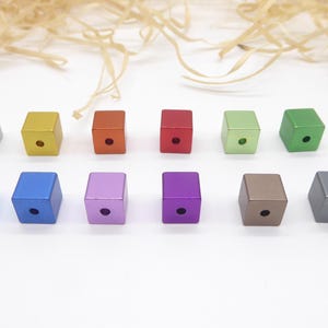 May include: Twelve colourful, square-shaped metal beads are displayed. The beads are in various colours, including silver, gold, orange, red, green, blue, purple, and brown. Each bead has a small hole in the centre. The beads are arranged on a white surface.