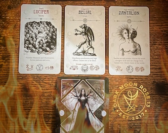 May include: A tarot card reading with cards featuring illustrations and text. The cards include "Lucifer," "Belial," "Dantalion," and "Revenge." The image also shows a deck of cards with a red symbol and two decorative stones. The Dark Moon Rose 13 logo is visible.