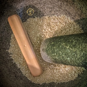 May include: A close-up of a mortar and pestle with a wooden pestle and a green mortar. The mortar is filled with white, flaked material.