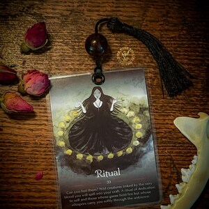 May include: A tarot card with a dark background and a woman in a black dress standing in a circle of yellow lights. The card is titled "Ritual" and has the number 33 on it. The text on the card reads: "Can you feel them? Wild creatures linked by the very blood you will spill into your craft. A ritual of dedication to self and those whose gazes hold fire but whose whispers carry you softly through the unknown."