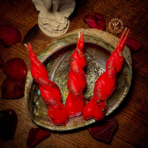 May include: Three red twisted candles in a seashell dish. The candles are decorated with glitter and have a rustic look. The dish is on a wooden surface with rose petals scattered around it.