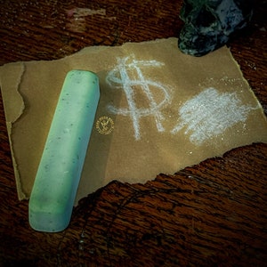 May include: A green chalk stick lies on a brown paper with a dollar sign drawn on it. A white powder is scattered around the dollar sign. A black skull sits on the paper.