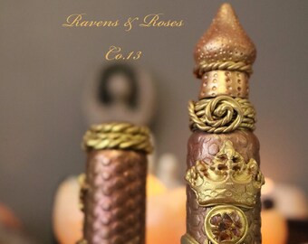 May include: Two gold and brown glass bottles with intricate details. The larger bottle has a gold bee, crown, and gemstone design. The smaller bottle has a gold rope design. The bottles are labeled "Ravens & Roses Co.1.3".