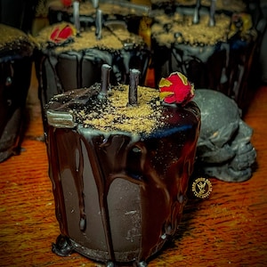 May include: Black candle with a dried red rose on top, surrounded by other black candles. The candle is dripping wax and has a brown, grainy texture. The candles are on a wooden surface.