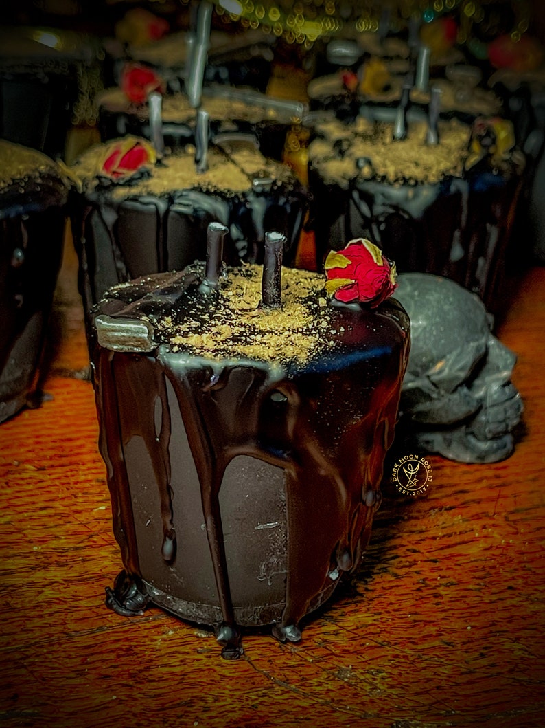 May include: Black candle with a dried red rose on top, surrounded by other black candles. The candle is dripping wax and has a brown, grainy texture. The candles are on a wooden surface.
