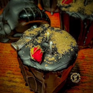 May include: Black candle with a red rose and gold flakes on top. The candle is dripping wax and has a label that says "Dark Moon Rose Est. 2017".