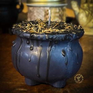 May include: Black cauldron candle with a single wick and dried herbs on top. The candle is dripping wax and has a dark moon rose logo on the side.