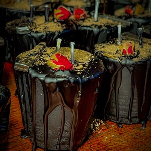 May include: Close-up of a dark chocolate candle with a red rose on top. The candle is drizzled with chocolate and has a gold glitter finish. The candle is labeled "Dark Moon Rose" and "Est. 2017".