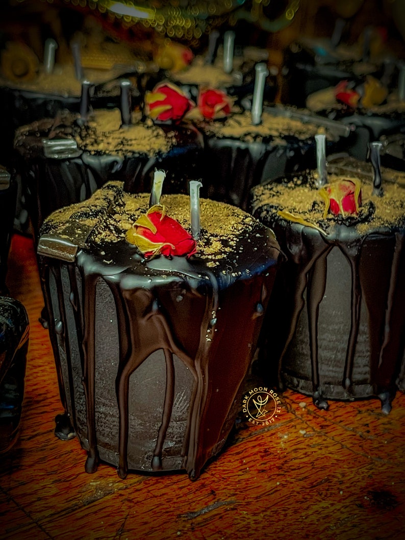 May include: Close-up of a dark chocolate candle with a red rose on top. The candle is drizzled with chocolate and has a gold glitter finish. The candle is labeled "Dark Moon Rose" and "Est. 2017".