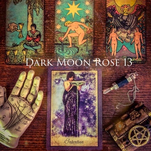 Op de afbeelding: Een assortiment tarotkaarten, een hand van handlezen, een pendel en een decoratieve doos zijn gerangschikt op een houten oppervlak. De kaarten bevatten illustraties van figuren en symbolen. De tekst "DARK MOON ROSE 13" en "Intention" zijn zichtbaar.