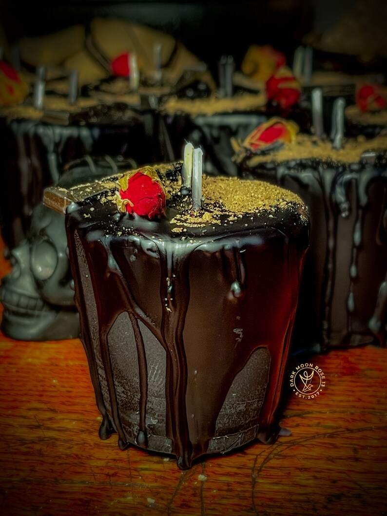 May include: A black candle with a red rose on top and gold glitter. The candle is dripping wax and has a label that says "Dark Moon Rose Est. 2017".