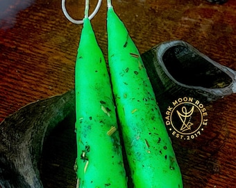 May include: Two bright green candles with a white wick loop, adorned with dried herbs. The candles are on a dark wooden surface, with a partial view of a skull and the text "DARK MOON ROSE 13 EST.2017".