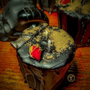 May include: Black candle with gold flakes and a red rose on top. The candle is dripping wax and has a gold label that says "Dark Moon Rose 13 Est. 2017".