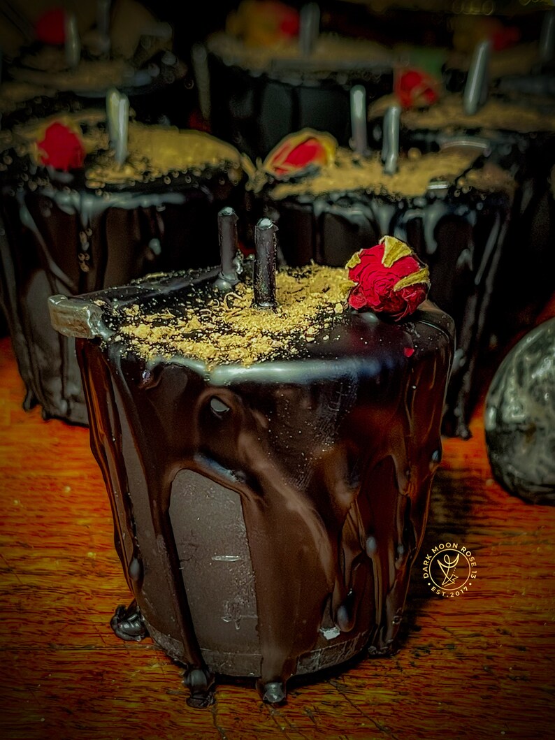 May include: A black candle with a dark chocolate coating and a dried red rose on top. The candle is in a glass jar and has a gold label with the text "Dark Moon Rose 13 Est. 2017".