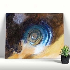 Könnte beinhalten: Leinwanddruck mit einem Satellitenbild der Richat-Struktur in Mauretanien. Das Bild zeigt eine kreisförmige geologische Formation mit Blau-, Braun- und Gelbtönen. Das Kunstwerk wird an einer weißen Wand neben einer Topf-Sukkulente ausgestellt.