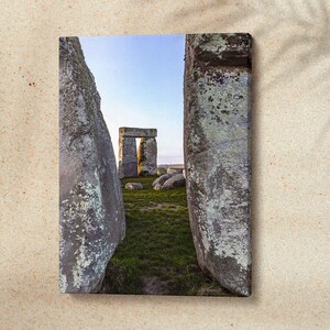May include: A canvas print featuring Stonehenge, a prehistoric monument. The image showcases the iconic stone circle, framed by two large, weathered stones in the foreground. The sky is clear blue, and the grass is green. The overall scene evokes a sense of history and mystery.
