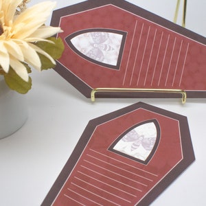May include: Two red coffin-shaped note cards with black trim and a white moth graphic in a window. The cards are on a gold stand.