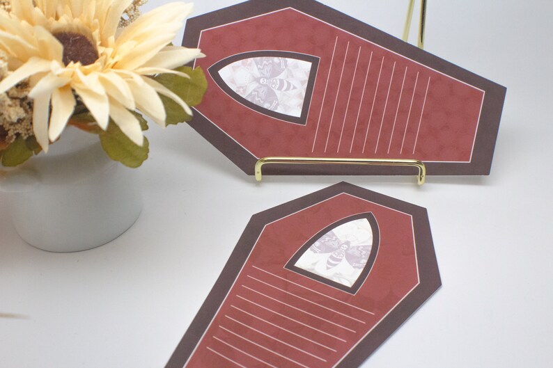May include: Two red coffin-shaped note cards with black trim and a white moth graphic in a window. The cards are on a gold stand.