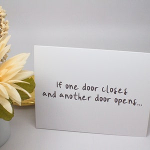 May include: A white card with the text "If one door closes and another door opens..." written in black ink. The card is sitting on a white surface next to a bouquet of cream-colored flowers.