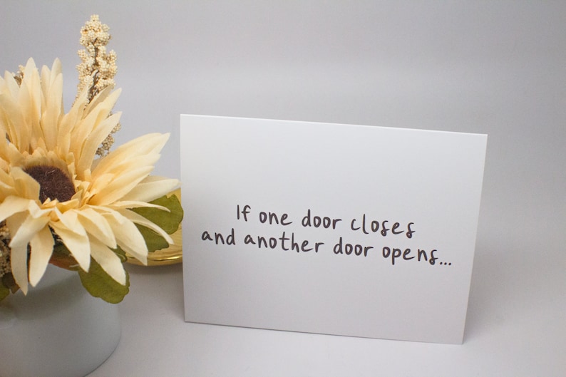 May include: A white card with the text "If one door closes and another door opens..." written in black ink. The card is sitting on a white surface next to a bouquet of cream-colored flowers.