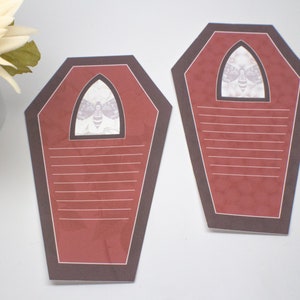 May include: Two red and brown coffin-shaped note cards with a white moth graphic in the top window. The cards have white lines for writing.
