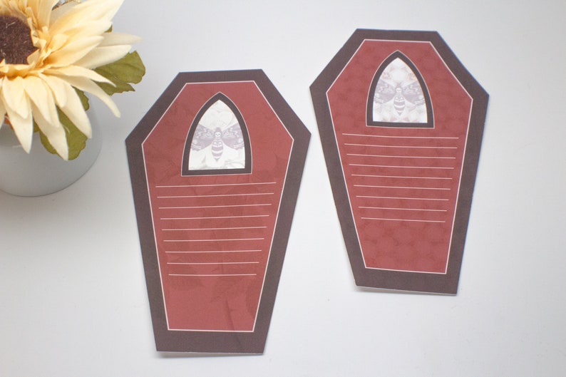 May include: Two red and brown coffin-shaped note cards with a white moth graphic in the top window. The cards have white lines for writing.