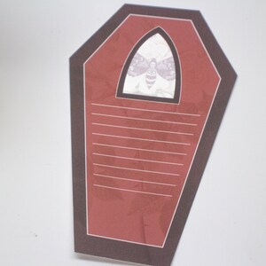 May include: A red coffin-shaped notepad with a black border and a white moth illustration in the top center. The notepad has lined paper for writing.