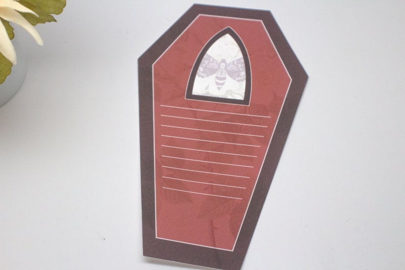 May include: A red coffin-shaped notepad with a black border and a white moth illustration in the top center. The notepad has lined paper for writing.