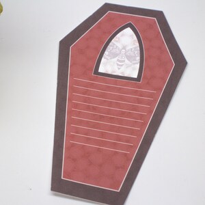 May include: A red coffin-shaped card with a black border and a white window featuring a black and white moth illustration. The card has lined writing space inside.