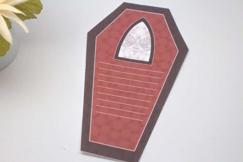 May include: A red coffin-shaped card with a black border and a white window featuring a black and white moth illustration. The card has lined writing space inside.
