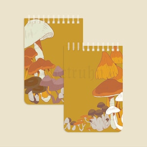 Puede incluir: Dos cuadernos con espiral con fondo amarillo mostaza. Cada cuaderno presenta una colorida ilustración de varias setas en tonos marrón, naranja y blanco. Los cuadernos están diseñados con un tema caprichoso inspirado en la naturaleza.