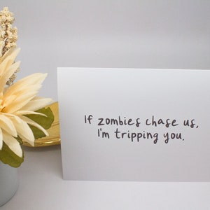 May include: A white greeting card with the text "If zombies chase us, I'm tripping you." is displayed. A decorative flower arrangement with a cream-colored flower and green leaves is in the background.