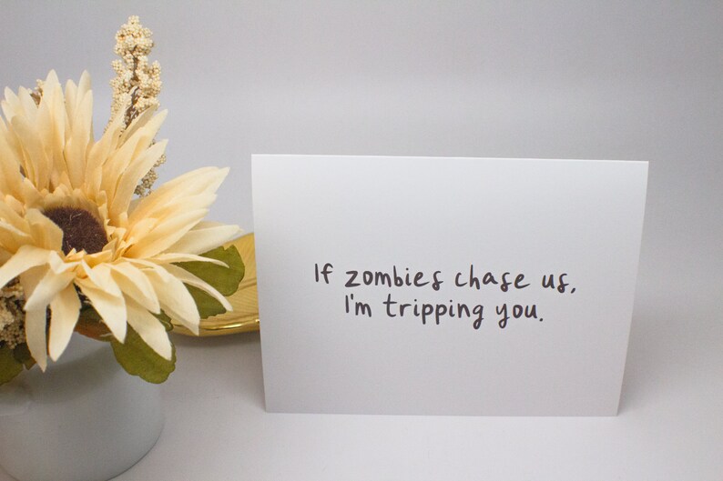 May include: A white greeting card with the text "If zombies chase us, I'm tripping you." is displayed. A decorative flower arrangement with a cream-colored flower and green leaves is in the background.