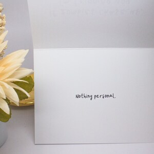 May include: A white greeting card with the phrase "Nothing personal." written in black script. The card is open, revealing the inside. A decorative flower arrangement with cream and brown tones is visible on the left.
