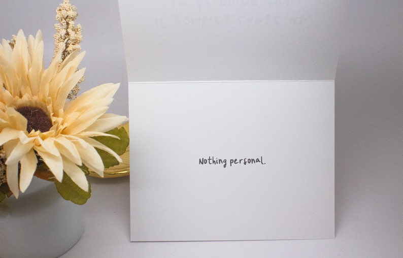 May include: A white greeting card with the phrase "Nothing personal." written in black script. The card is open, revealing the inside. A decorative flower arrangement with cream and brown tones is visible on the left.