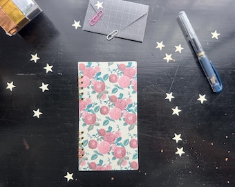 May include: A floral-patterned notebook with pink roses and green leaves lies on a dark surface, next to a pen, a small envelope with a pink paperclip, and star-shaped confetti. The notebook has a spiral binding.