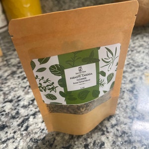 May include: A brown paper bag with a white label that reads "Parasite-Candida Cleanse loose herbal blend 1 oz" and features a green leafy design. The bag contains dried herbs.