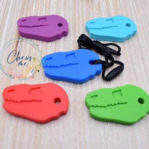 May include: Five colorful dinosaur-shaped chewable pendants in purple, blue, red, green, and turquoise. One blue pendant is attached to a black cord. The pendants are designed for sensory needs and feature a textured surface.