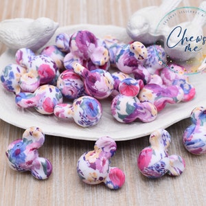 May include: A collection of floral patterned silicone mouse ears. The ears are in a white bowl on a wooden surface. The ears are a light purple, pink, and blue color with a white background.