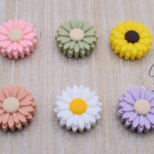 May include: Six colorful daisy-shaped silicone beads in pink, green, yellow, peach, white, and lavender. Each bead has a textured petal design and a contrasting center. The beads are arranged on a light wood-grain surface.
