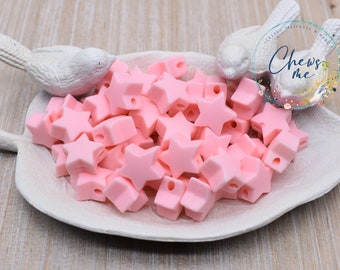 May include: A pile of pink silicone star-shaped beads with holes for threading. The beads are in a white ceramic dish with two white bird figurines on either side. The text "Chews Me" is visible in the background.