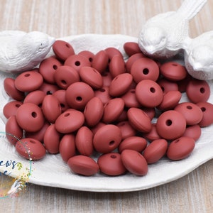 May include: A close-up of a pile of 25mm silicone beads in a deep burgundy color. The beads have a hole in the center and are arranged in a bowl.
