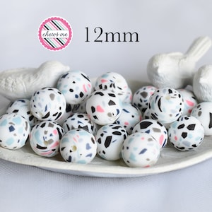 May include: A close-up of a bowl filled with 12mm silicone beads. The beads are white with a terrazzo pattern in black, pink, and blue. The beads are 12mm in diameter.